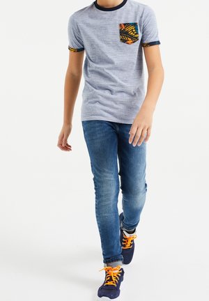 Junge läuft und trägt ein hellgraues T-Shirt mit gemustertem Taschen- und Ärmeldesign, blaue Jeans und marineblaue Sneaker mit leuchtend orangen Schnürsenkeln.