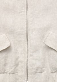 Veste en lin beige avec une fermeture éclair frontale, deux poches plaquées et un détail froncé à la taille pour une silhouette ajustée.