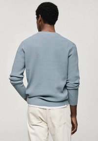 Homme vu de dos portant un pull côtelé bleu clair et un pantalon blanc cassé, les mains partiellement dans les poches, devant un fond uni.