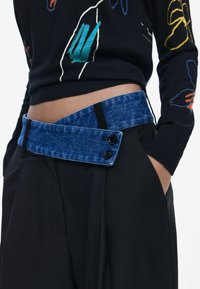 Jeansgürtel mit blauer Textur und zwei schwarzen Knöpfen, getragen über schwarzen Hosen mit Seitentaschen. Schwarzer Pullover mit buntem Blumenmuster.