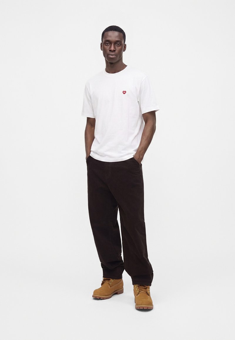 Ein Mann steht mit den Händen in den Taschen, trägt ein weißes T-Shirt mit kleinem rotem Herz, schwarze Hose und beige Stiefel vor einfachem weißem Hintergrund.