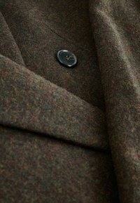 Uldblanding i nuancer af mørkegrøn og brun, der har en stor sort knap og et struktureret, foldet design med synlige sømme.