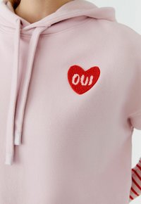 Sweat à capuche rose en coton, avec un cœur rouge brodé et le texte "OUi". Texture lisse, avec cordons de serrage et poignets côtelés.
