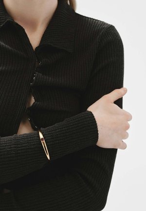 Suéter negro de canalé con botones, de manga larga, combinado con una pulsera dorada que presenta un acento de gema roja en la muñeca.