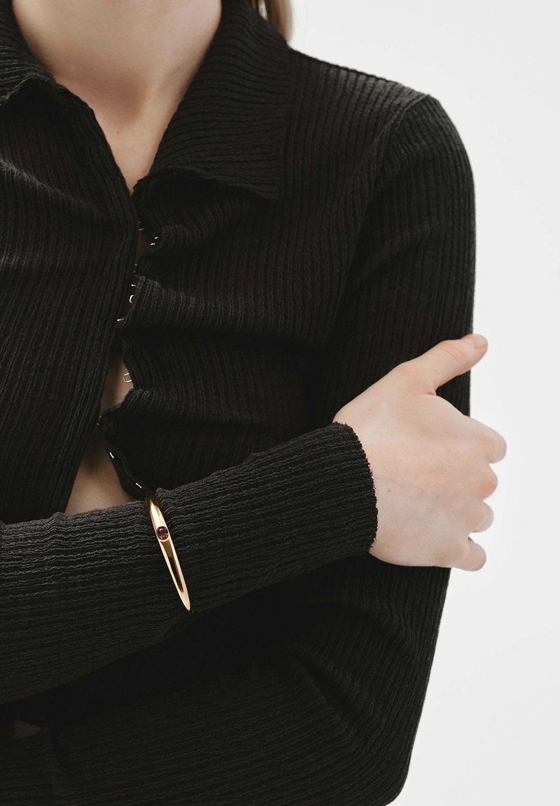 Suéter negro de canalé con botones, de manga larga, combinado con una pulsera dorada que presenta un acento de gema roja en la muñeca.