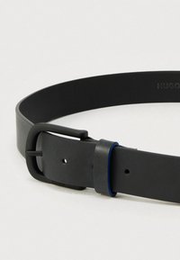 Juodas odinis diržas su matinio paviršiaus apdaila, turi stačiakampį juodą sagtį ir mėlyną akcentą ant segtuko kilpos. Lygus paviršius.