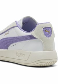Sneaker Puma cu un superior din piele albă, cu accente din suede lavandă, un toc texturat și o talpă din cauciuc. Detaliu cu logo auriu pe lateral.