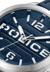 Marinblå klocka med en texturerad urtavla, djärva vita siffror, silverfärgade visare och ett hölje i rostfritt stål. Läderremmens detaljer synliga.