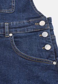 Jeansoverall med en ficka med gul söm och silverfärgade metallknappar. Tyget är mörkblått med en strukturerad yta.