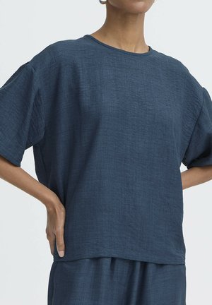 Person iført løstsiddende, tekstureret mørkeblå kortærmet top og matchende bukser, stående med hænderne på hoften foran en ensfarvet baggrund.