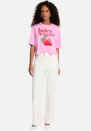 Femme aux cheveux bouclés portant un t-shirt rose avec l'inscription "Juicy & Sweet" et un motif fraise, un pantalon blanc taille haute et des talons blancs.