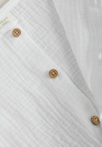 Vêtement de bébé blanc texturé avec des boutons en bois étiquetés "NEXT" et une étiquette en tissu indiquant "NEXT baby."