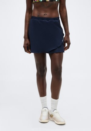 Žena má na sebe tmavomodrý športový skort, biele ponožky a biele tenisky, stojí pred jednoduchým svetlým pozadím.