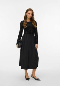 Melnā garo piedurkņu blūze ar teksturētu rakstu, saskan ar plūstošu melnu svārku. Aksesuāri ietver mazu melnu somiņu un garus, norādītus zābakus.