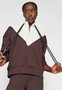 Brun og kremfarget fargeblokkert sweatshirt med glidelås i halsen, med en trekantet design og ribbestrikkede mansjetter. Modellen holder svarte motstandsbånd.