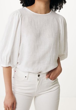 Mujer con blusa de lino blanca con mangas abullonadas y pantalones blancos de cintura alta, mano descansando en el bolsillo del pantalón.