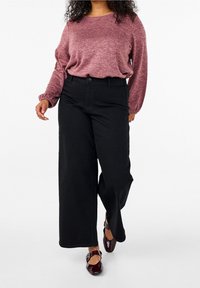 Donna che indossa un maglione rosa a maniche lunghe, pantaloni neri a gamba larga e scarpe Mary Jane marrone scuro, che cammina su uno sfondo bianco.