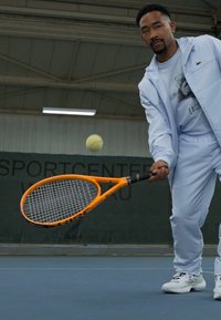 Tenisa rakete ar oranžu rāmi un melnu saķeri, ko tur personāžs gaiši zilos sporta tērpos, gatavojoties trāpīt tenisam.