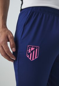 Tengerészkék sportnadrág elasztikus derékkal és oldalsó zsebekkel, rózsaszín hímzett pajzslogóval, csillagokkal és csíkkal díszítve.
