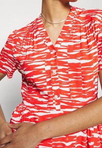 Kvinna i en röd och vit mönstrad knappad klänning med korta ärmar och ett silverhalsband, med armarna korsade vid midjan.