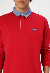 Red long-sleeve polo shirt with a light blue denim collar. Features a GANT logo patch on the chest and a button closure at the neck.