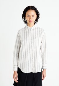 Chemise à manches longues à rayures en tissu léger. Présente des rayures verticales brunes sur fond blanc, un col et une poche poitrine unique.
