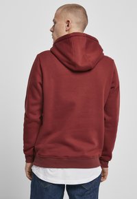 Sudadera burdeos con bolsillo tipo canguro, capucha ajustable con cordón y puños acanalados. Llevada sobre una camiseta blanca y combinada con jeans azules.