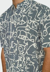 Korte ærmer button-up skjorte i blåt stof med et hvidt abstrakt ansigt og symbolmønster. Fremtrædende funktioner inkluderer en krave og en enkelt frontlomme.