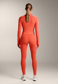 Hladký top s vysokým golierom a dlhými rukávmi a priliehavé legíny z jasne oranžového elastického materiálu, s minimálnym počtom švov a diskrétnym logom vzadu.
