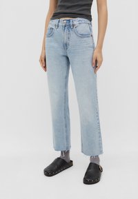 Jeans de pierna recta en azul claro con textura desgastada, dobladillo deshilachado, bolsillos frontales y cierre de botón, combinados con zuecos negros y calcetines grises.