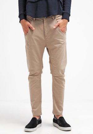 Persoon met beige slim-fit broek met handen in de voorzakken en zwarte instapschoenen, staand tegen een effen witte achtergrond.