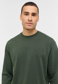 Žalias ilgarankovis džemperis su apvaliu reljefiniu logotipu ant krūtinės. Pagamintas iš minkštos medžiagos su tekstūruotu paviršiumi.