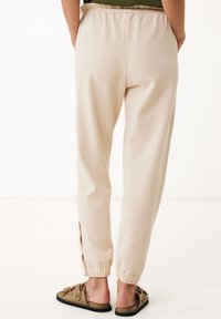 Beige joggers met elastische tailleband en taps toelopende boorden. Glad materiaal met zijsplitten, gecombineerd met bruine instapsandalen met een decoratief logo.