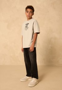 Maglietta beige oversize con motivo grafico nero, abbinata a jeans in denim scuro e sneaker chiare. Tessuto liscio, vestibilità comoda.