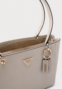 Beige strukturierte Handtasche mit goldfarbenen Beschlägen, ausgestattet mit Reißverschluss, zwei Griffen und einem Markenzeichen in Dreiecksform. Mit Anhängern an der Tasche.