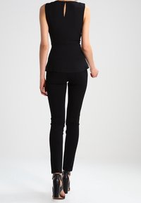 Top nero peplum con apertura a goccia sulla schiena, abbinato a pantaloni aderenti neri. L'insieme presenta texture lisce e scarpe eleganti con tacchi alti.