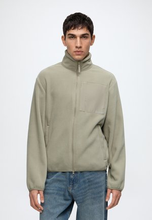 Mann trägt eine khakifarbene Fleecejacke mit Reißverschluss, hohem Kragen und Brusttasche, kombiniert mit blauen Jeans, stehend vor einfarbigem Hintergrund.
