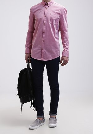 Mężczyzna w różowej koszuli na guziki, ciemnych spodniach i szarych butach wsuwanych, trzymający czarny plecak obok siebie na jednolitym tle.