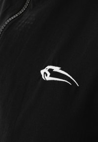 Weiß stilisierte Vogelstickerei auf schwarzem Stoff nahe einer Reißverschlussnaht, die auf ein Sport- oder Freizeitjackendesigndetail hinweist.