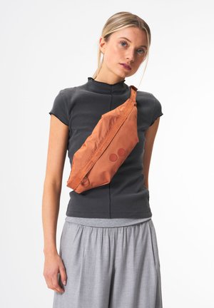 Sac en tissu orange en bandoulière avec une fermeture éclair diagonale, présentant deux accents circulaires. Porté en travers du corps, il a une texture lisse et une forme angulaire.