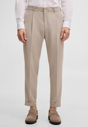 Man draagt beige geplooide broek met omslagen, bruine suède loafers en een witte blouse tegen een effen achtergrond.