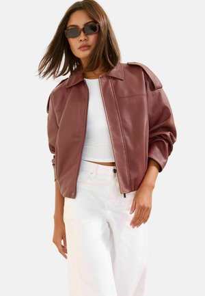 Žena v tmavočervenej koženej bunde, bielom crop tope, bielych nohaviciach a tmavých slnečných okuliaroch pózujúca na jednoduchom svetlom pozadí.