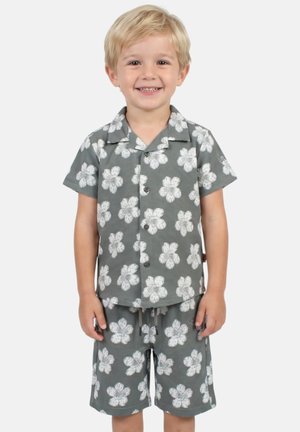 Jeune garçon souriant portant une chemise grise à manches courtes à boutons et un short assorti avec des motifs floraux blancs, debout devant un fond uni.