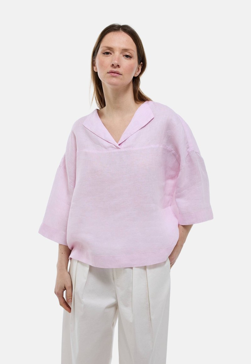 Blouse en lin rose clair avec un col en V, coupe ample et manches courtes. Présente une texture lisse et des fentes latérales pour le confort.