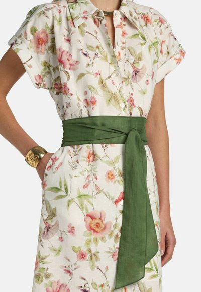 Vestido floral abotonado con flores rosas y verdes, mangas cortas, cinturón de tela verde atado a la cintura y pulsera de oro en la muñeca izquierda.