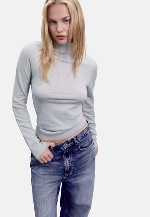 Hellgrauer, gerippter Rollkragenpullover mit langen Ärmeln, kombiniert mit hochtaillierten blauen Denim-Jeans. Der Stoff wirkt weich und figurbetont.