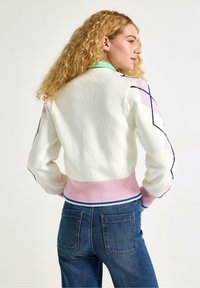 Pletemý bílý svetr s růžovými a modrými akcenty, s kontrastními vzory na rukávech a žebrovaným lemem. Nošeno s modrými džínami z denimu.