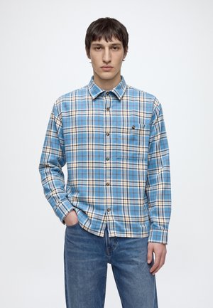 Un homme porte une chemise boutonnée à carreaux bleus et blancs avec un jean, sur un fond clair uni.