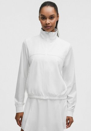 Vrouw die een witte jas met ritssluiting, hoge kraag en lange mouwen draagt, gecombineerd met een witte rok, staand tegen een effen achtergrond.