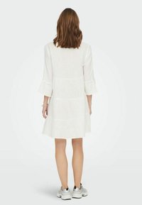 Weißes, knielanges Kleid mit weit ausgestellten Ärmeln, gestuftem Design und strukturiertem Stoff. Getragen über Sneakers für einen lässigen Look.
